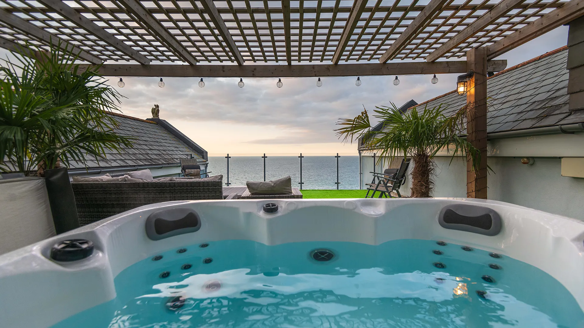 A luxurious outdoor hot tub with bubbling water, set under a wooden pergola with string lights, overlooking the sea at sunset.