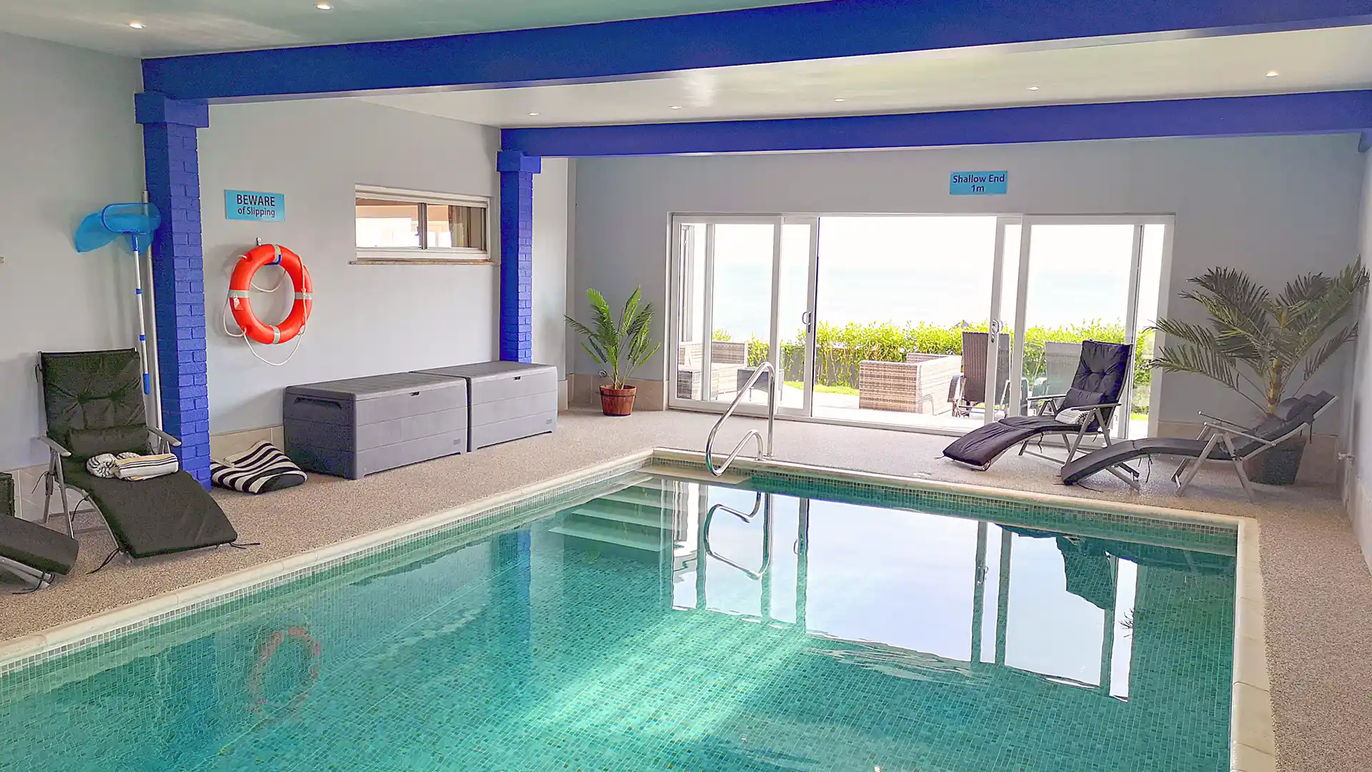 Indoor swimming pool with turquoise mosaic tiles, surrounded by lounge chairs and tropical plants, with glass doors leading to an outdoor seating area and sea views.