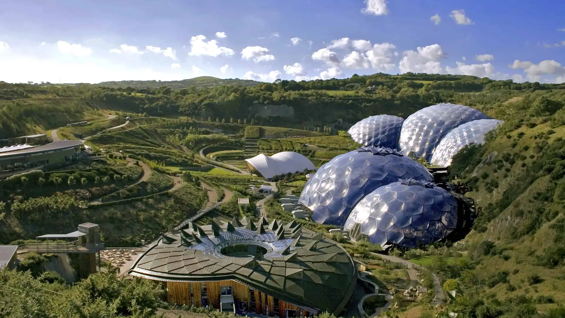 A panoramic view of the Eden Project in Cornwall, showcasing its iconic biomes nestled within lush, rolling hills. The geometric domes glisten under the sunlight, creating a futuristic contrast against the natural landscape.