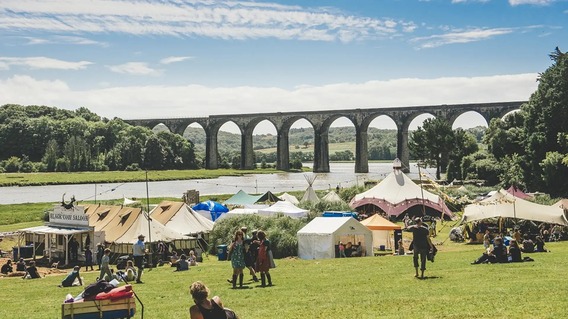 Festival scene on a sunny day with a variety of tents and marquees set up in a lush green field, with a large viaduct spanning across the background.