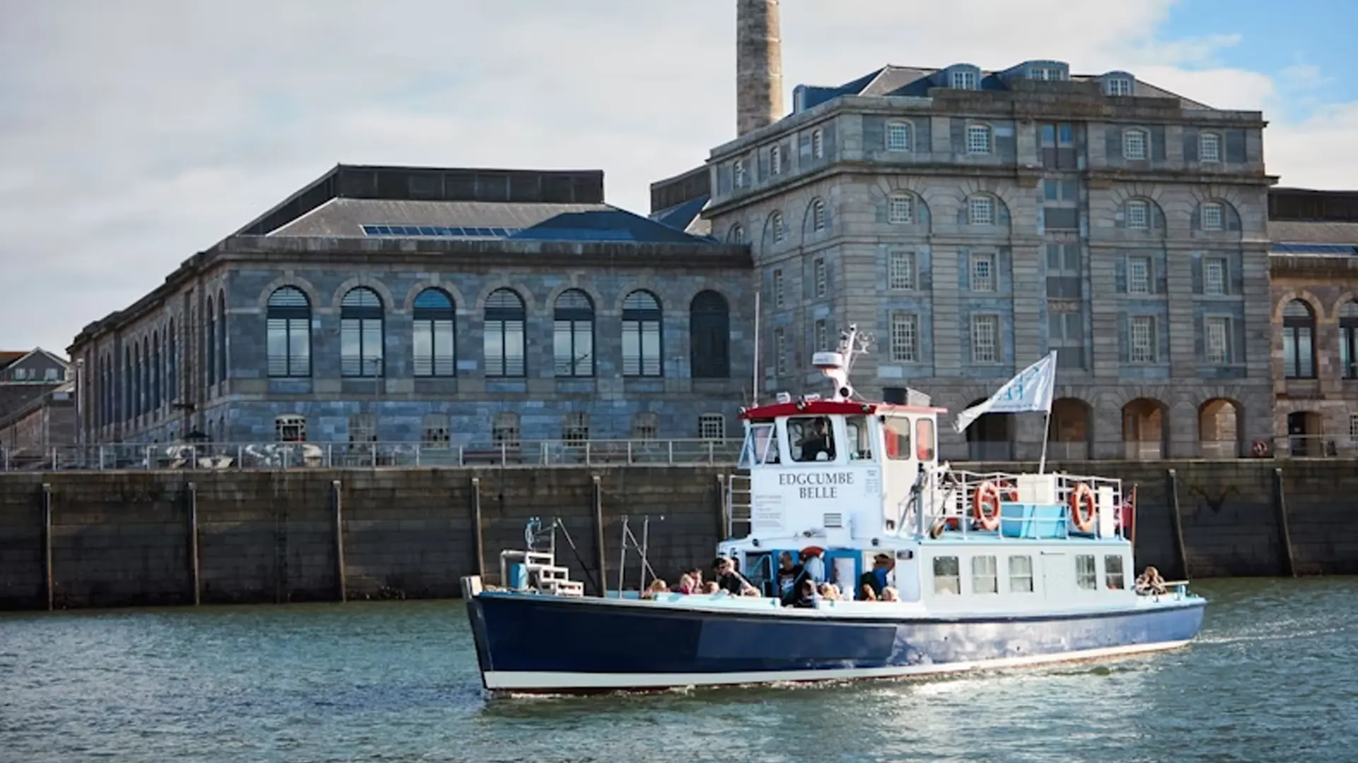 A blue and white passenger ferry named 'Edgcumbe Belle' glides through the water with several passengers on board, against the backdrop of a grand historic waterfront building featuring arched windows and stone architecture. The ferry's flag flutters in the breeze, adding to the maritime charm of the scene.