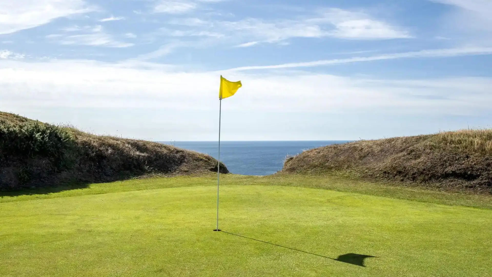 A golf green with a yellow flag set against a backdrop of rolling dunes and the open sea under a bright blue sky.