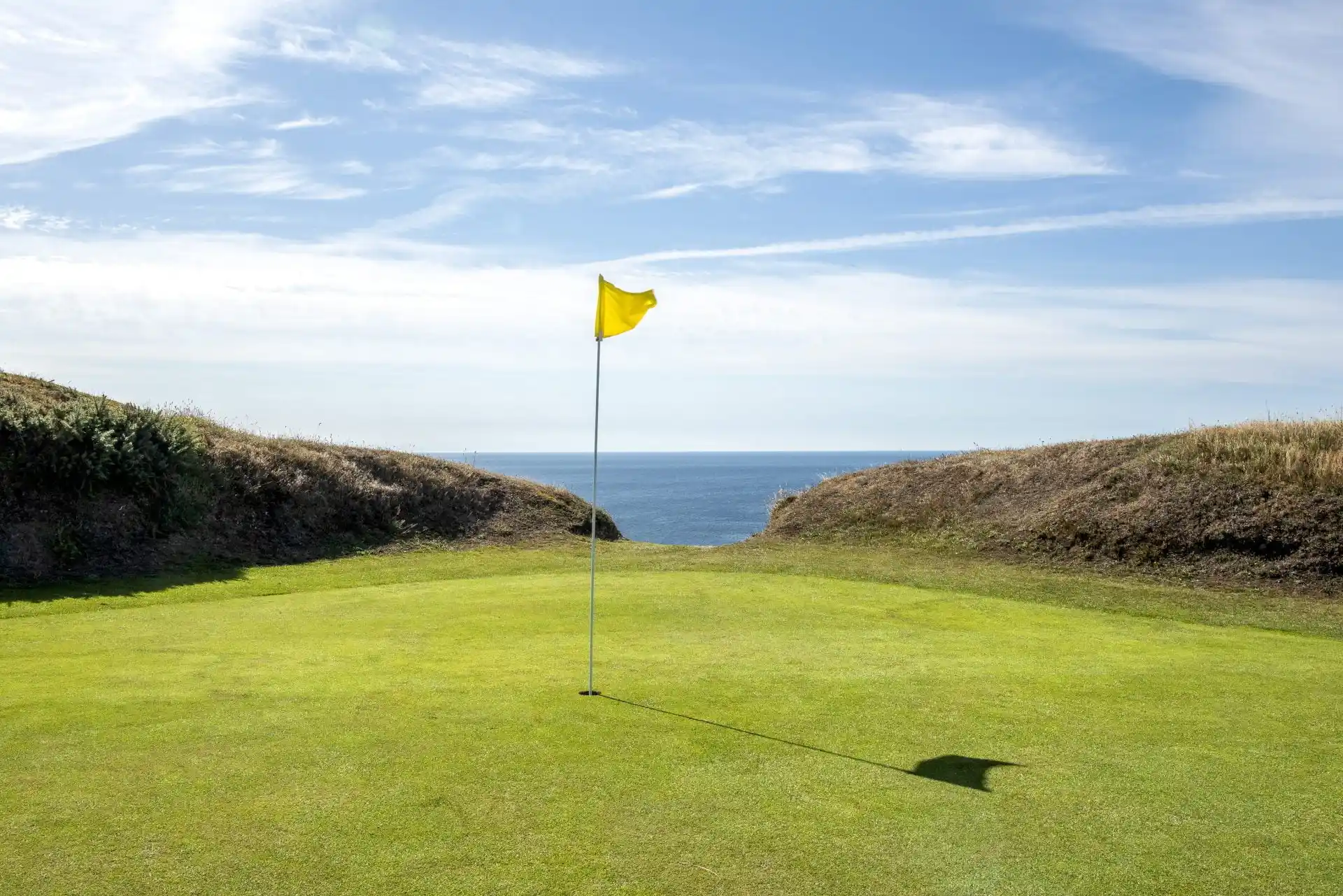A golf green with a yellow flag standing prominently, set against a backdrop of rolling dunes and a clear ocean view.